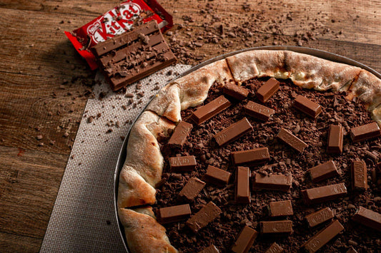 Chocolate pizza topped with KitKat pieces on a rustic wooden table, with a partially opened KitKat wrapper beside it.