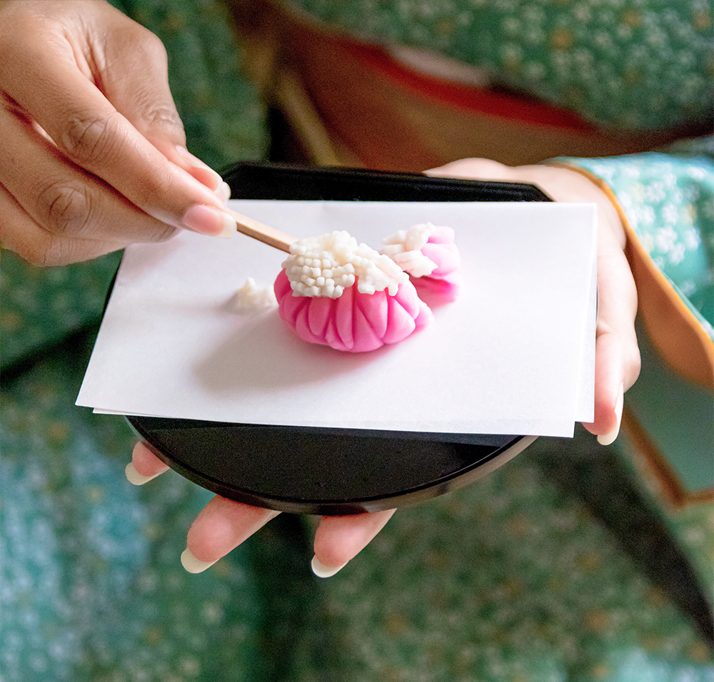 Person in traditional attire holding a pink Japanese wagashi dessert with chopsticks on a lacquer dish.